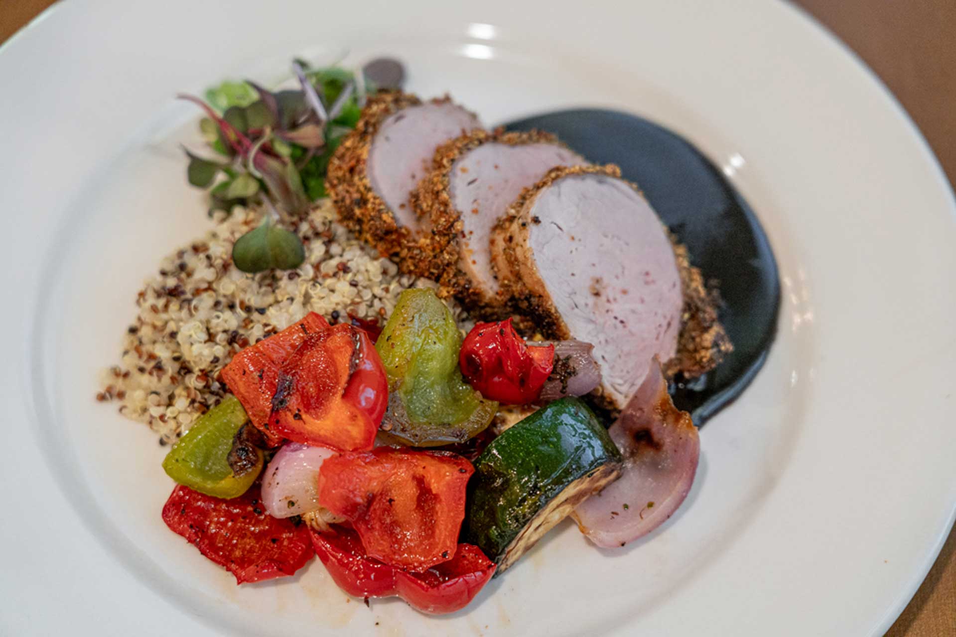 A plated meal at MorningStar Senior Living Hillsboro features sliced pork tenderloin with a dark sauce, quinoa, roasted peppers, zucchini, onions, and microgreens—a delicious option for those enjoying vibrant Retirement Living.