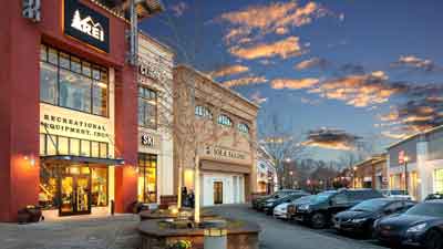 Outdoor shopping center at sunset with a REI store on the left, other retail shops, parked cars, and a tree in the foreground under a partly cloudy sky—just minutes from MorningStar Senior Living Hillsboro and ideal for Retirement Living convenience.