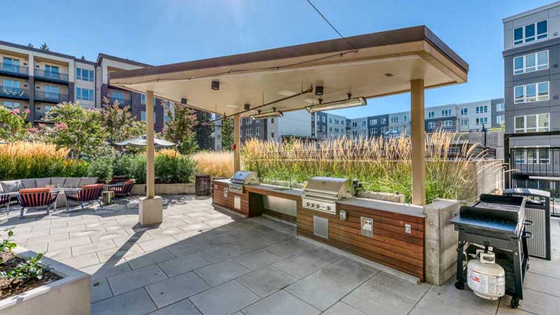 Modern outdoor grilling area with covered barbecue stations, surrounded by tall grasses and seating, set in a landscaped courtyard of MorningStar Senior Living Hillsboro, perfect for retirement living on a sunny day.