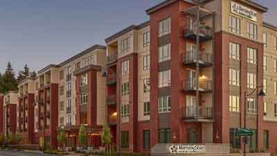 Modern multi-story apartment building with brick and beige siding, balconies, large windows, and landscaped greenery in front. The “Merrill Gardens” sign highlights comfortable retirement living beneath a clear sky.