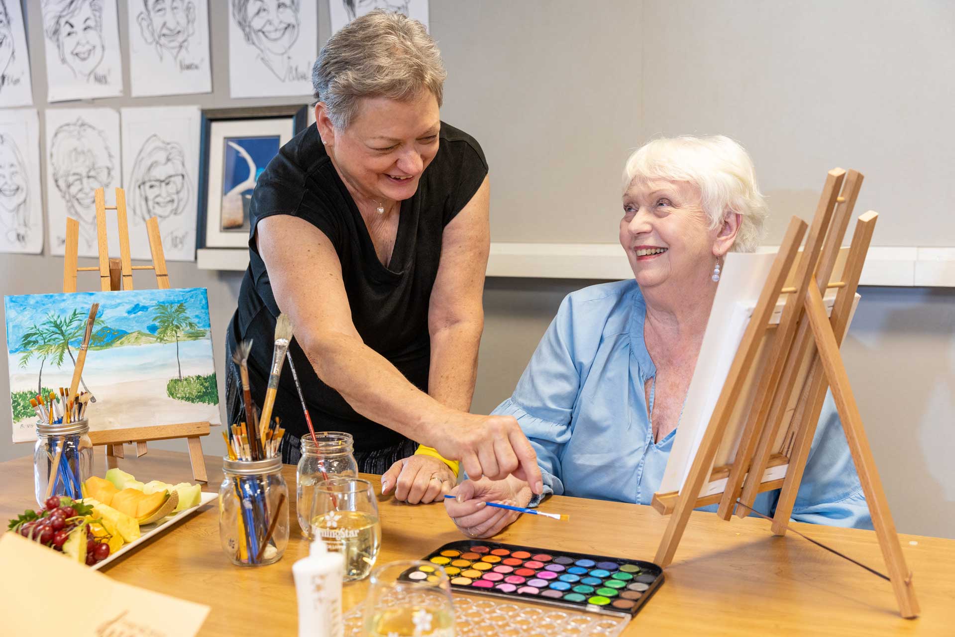 Two older women at MorningStar Senior Living Hillsboro enjoy a painting activity together, smiling as one helps the other with her brush. Art supplies and watercolors fill the table, reflecting the joy of creative retirement living.