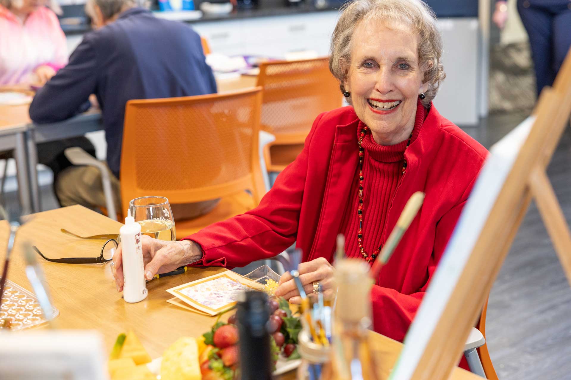 An elderly woman in a red jacket smiles while sitting at a table with art supplies, a candle, and snacks in the brightly lit community room at MorningStar Senior Living Hillsboro, enjoying retirement living with others in orange chairs nearby.