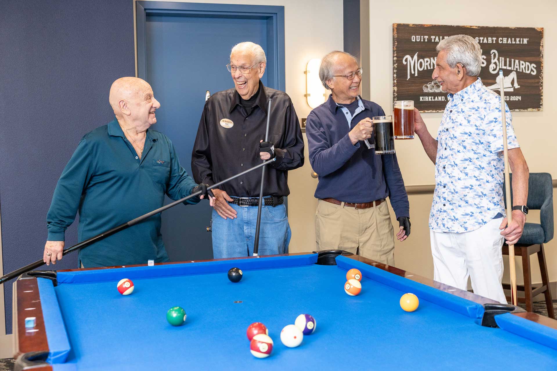Four older men stand around a pool table at MorningStar Senior Living Hillsboro, smiling and enjoying each other's company. One holds a cue stick, another has a large mug of beer. A billiards sign hangs on the wall—true retirement living joy.