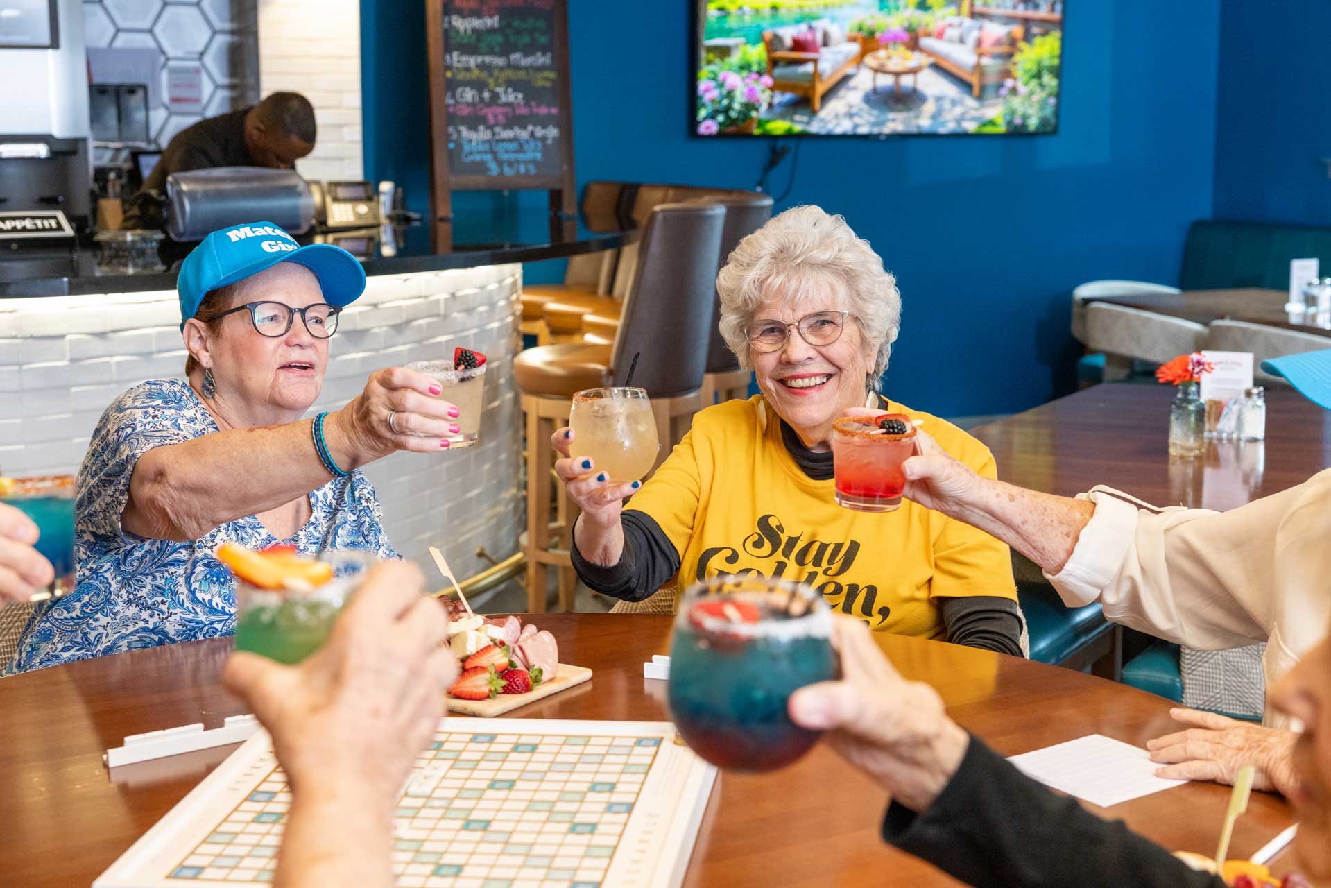 A group of older adults at MorningStar Senior Living Hillsboro sits around a table, smiling and raising colorful drinks in a toast. Scrabble and snacks add to the cheerful, relaxed atmosphere in this bright, modern retirement living space.