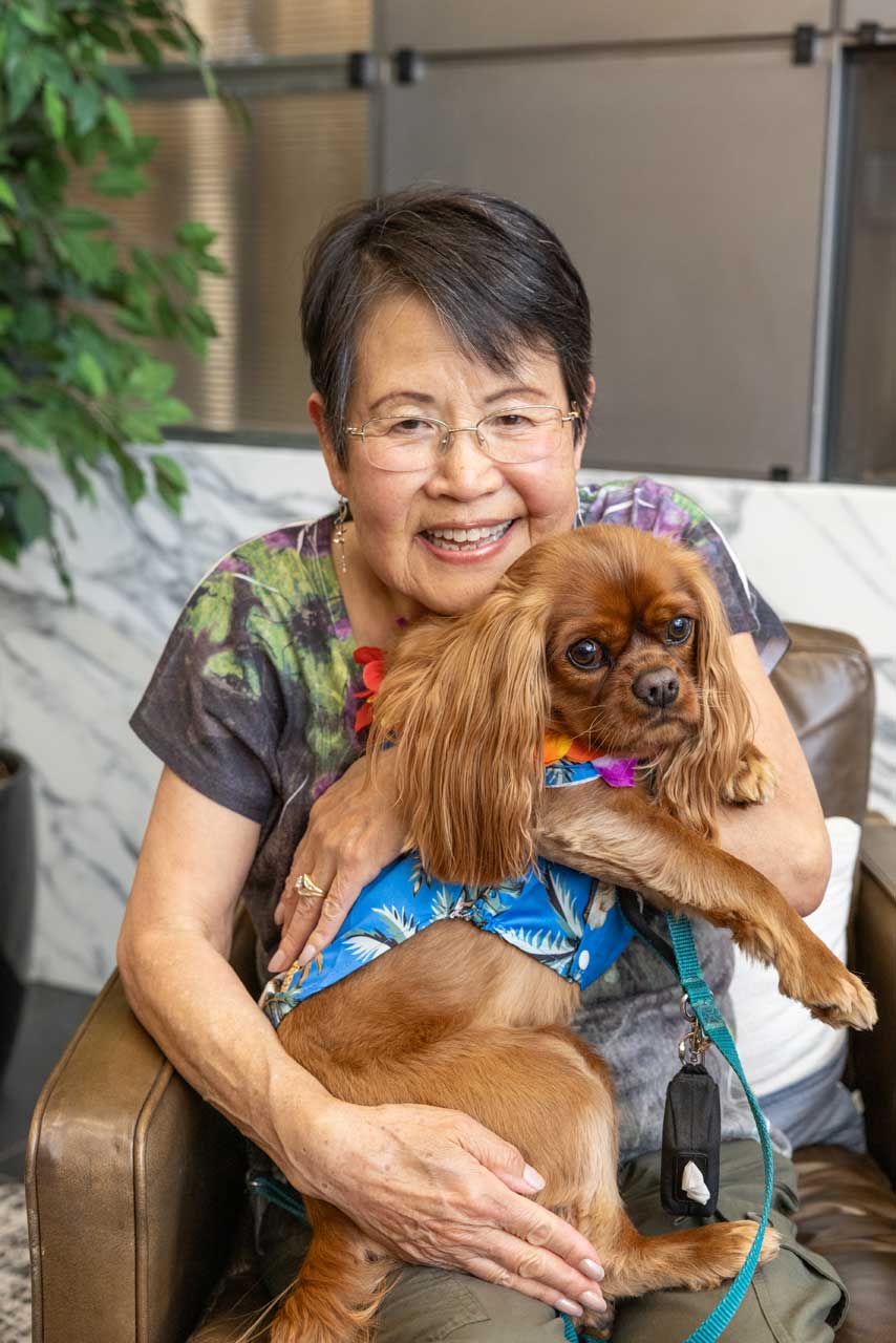 An older woman with short dark hair and glasses smiles while hugging a brown Cavalier King Charles Spaniel in a blue harness, sitting together on a chair indoors at MorningStar Senior Living Hillsboro, enjoying the warmth of retirement living.