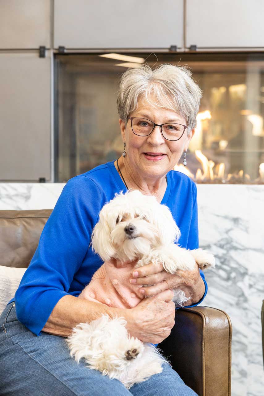 An older woman with short gray hair and glasses sits on a couch at MorningStar Senior Living Hillsboro, smiling as she holds a small white fluffy dog in a pink sweater. A modern fireplace is in the background, reflecting the warmth of retirement living.