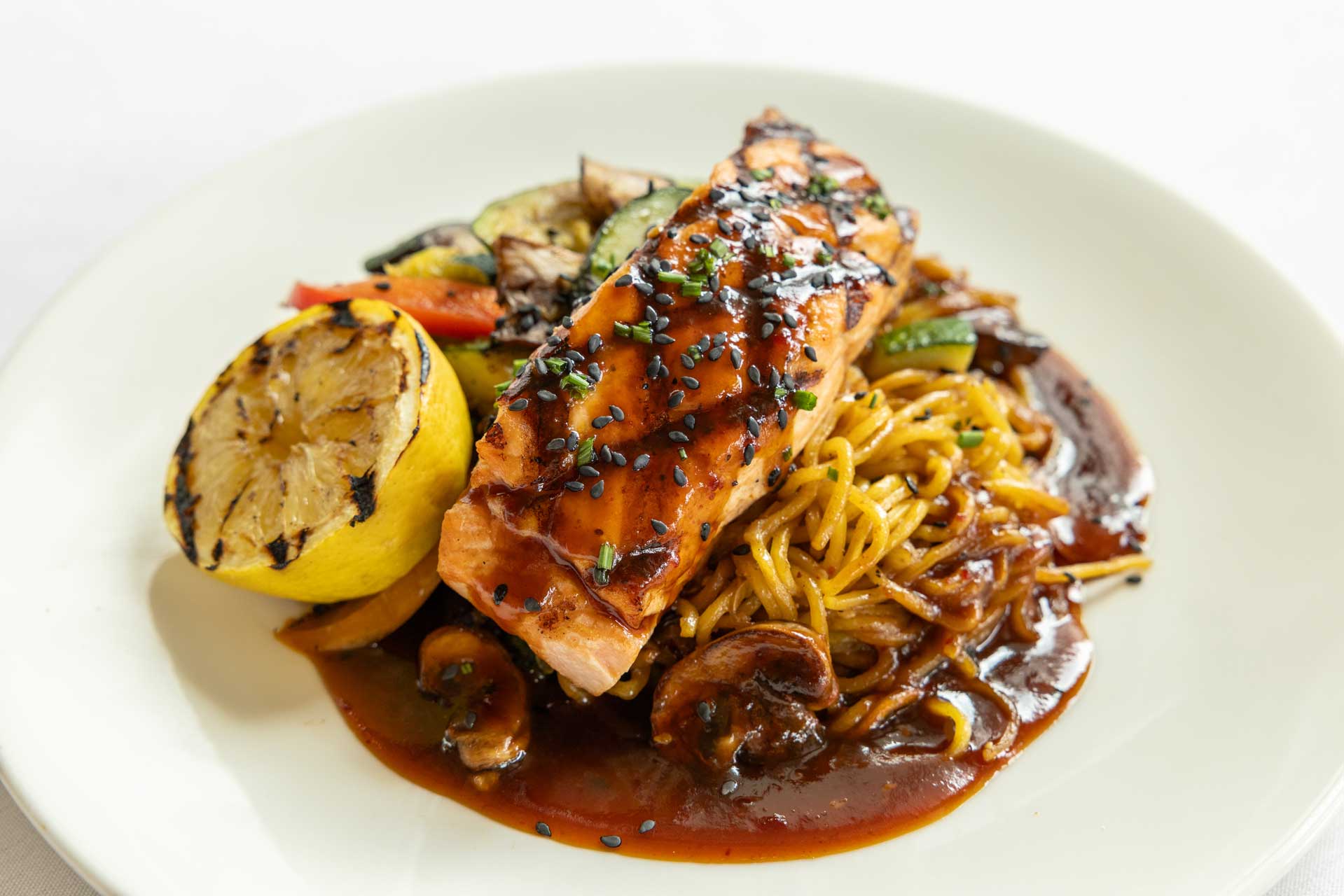 Grilled salmon with black sesame seeds, served over noodles and mushrooms in a brown sauce, accompanied by grilled vegetables and a charred lemon half—a delicious option at MorningStar Senior Living Hillsboro, redefining retirement living.