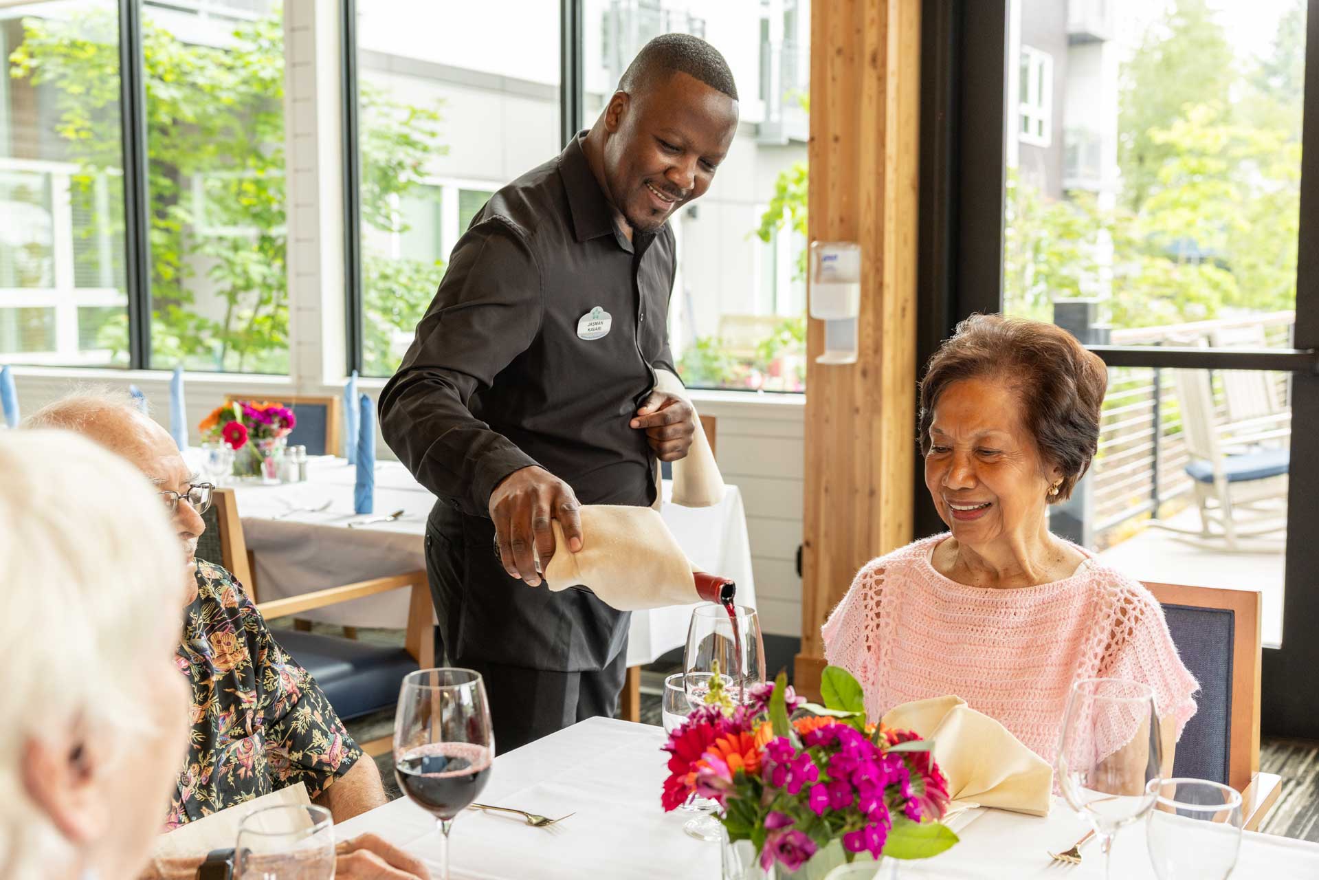 A smiling waiter pours red wine for an elderly woman seated with others in the bright, modern dining room at MorningStar Senior Living Hillsboro, where retirement living is enjoyed amid large windows and fresh flowers.