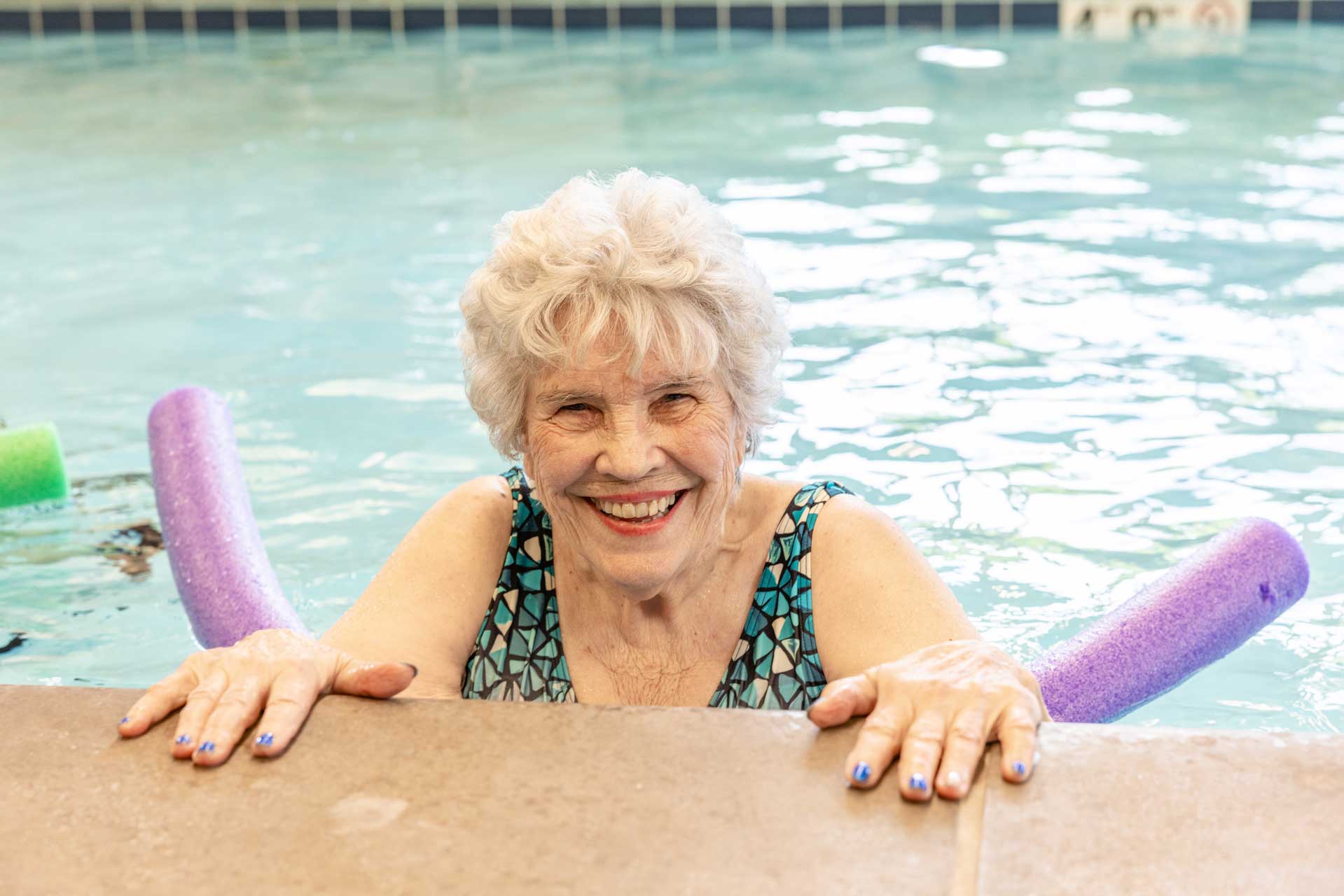 An elderly woman with short white hair smiles while holding the edge of an indoor pool at MorningStar Senior Living Hillsboro. She wears a patterned swimsuit, colorful pool noodles floating behind her, enjoying vibrant retirement living.