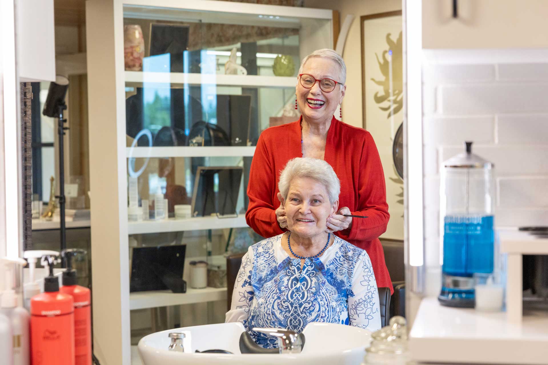In a bright, modern salon at MorningStar Senior Living Hillsboro, two elderly women share a joyful moment—one styles the other's hair as they both smile, surrounded by salon products and a mirror, reflecting the warmth of retirement living.