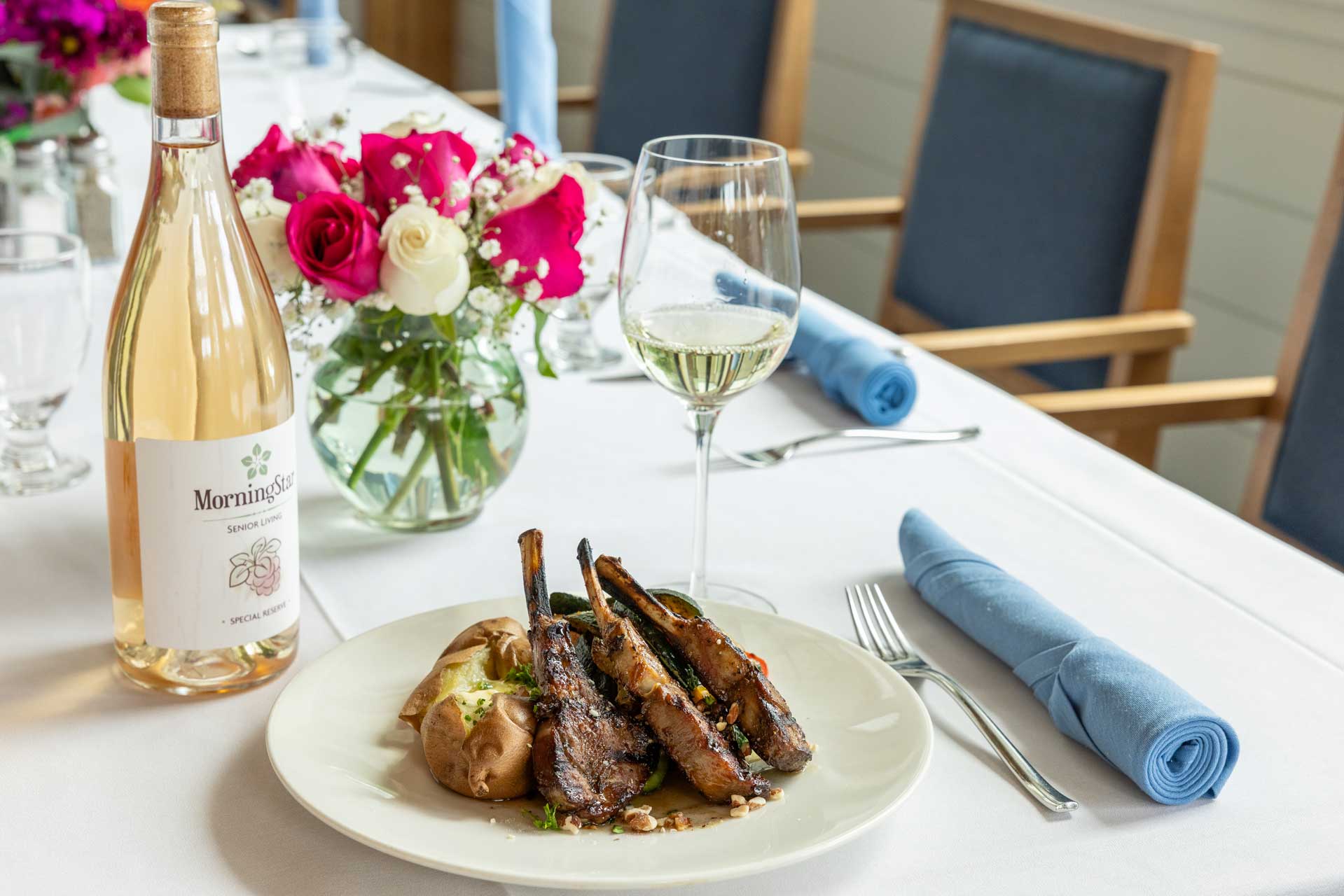 A plate of grilled lamb chops with a baked potato sits on a white tablecloth next to a glass of white wine, a bottle of rosé, and a vase of pink and white flowers—fine dining at MorningStar Senior Living Hillsboro. Blue napkins are neatly rolled beside the plate.