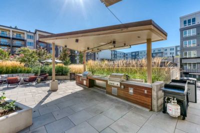 Outdoor patio area at MorningStar Senior Living Hillsboro features a covered grilling station, built-in barbecue grills, and a cozy fire pit—perfect for enjoying retirement living amid tall grasses and modern apartments under a clear blue sky.