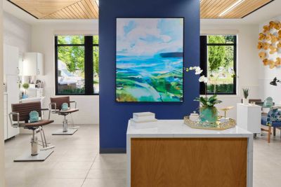 Modern salon interior at MorningStar Senior Living Hillsboro with styling chairs, large abstract artwork on a blue accent wall, white counters, and windows letting in natural light. Decor includes plants and yellow wall art on the right.
