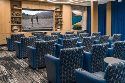 Cozy theater room at MorningStar Senior Living Hillsboro, featuring patterned blue chairs arranged in rows facing a large screen. Wood paneling, blue curtains, and wall art create a warm ambiance ideal for retirement living.