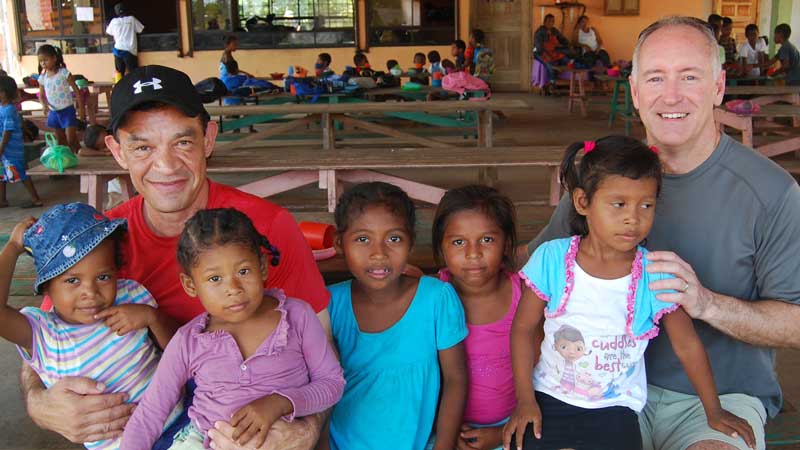 Two men sit with five young girls on their laps and by their sides, all smiling for the camera. In the background at MorningStar Senior Living Hillsboro, other children and adults are seated at picnic tables inside a large, open building.