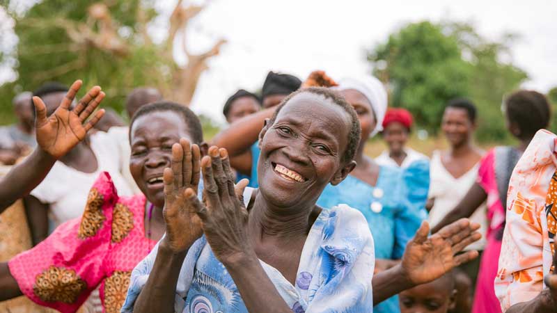 A group of women at MorningStar Senior Living Hillsboro enjoy retirement living outdoors, smiling, laughing, and clapping their hands in colorful clothing amid trees and greenery.