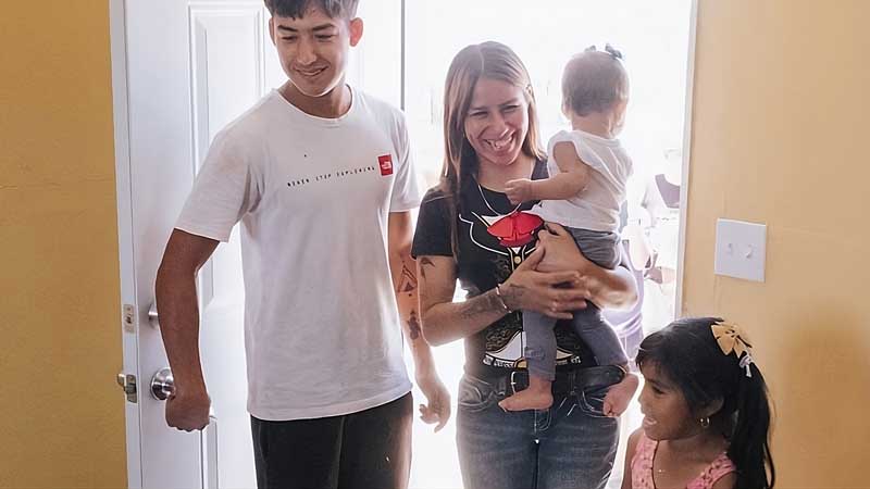 A smiling woman holding a baby stands in a doorway with a man and a young girl, all appearing happy as they enter a brightly lit room with yellow walls at MorningStar Senior Living Hillsboro.