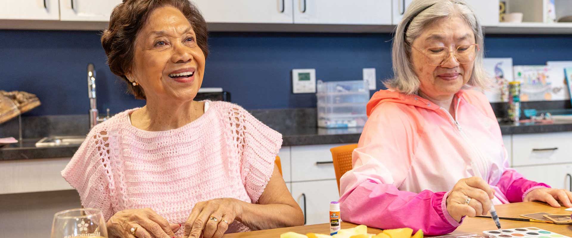 Two older women enjoy arts and crafts at MorningStar Senior Living Hillsboro. One smiles while folding paper; the other draws with a marker. A plate of fruit and cheese sits before them in a bright, cheerful retirement living space.