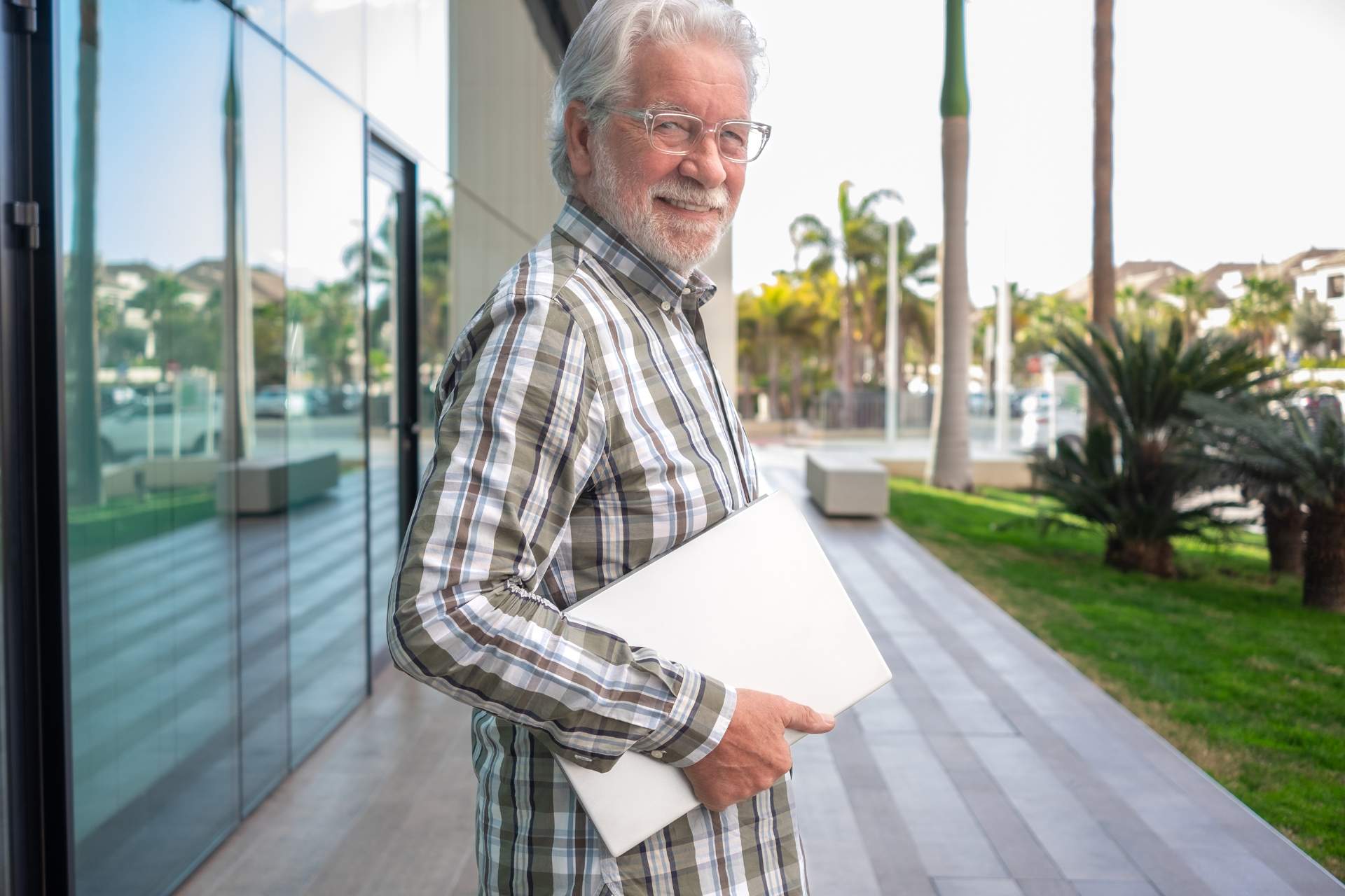 A senior man standing outside a building, holding a laptop and smiling